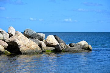 Scenic view of large natural rocks and boulders on a coastal shoreline with calm blue sea and clear sky in the background. Minimalist seascape symbolizing stability, nature, summer vacation.