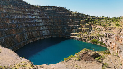 Aerial view of a deep blue crater lake of the Mary Kathleen Uranium Mine surrounded by steep rocky walls in a quarry. Turquoise water near the shore contrasts with the deep blue center