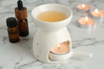 Aroma lamp with burning candles and essential oils on white marble table, closeup