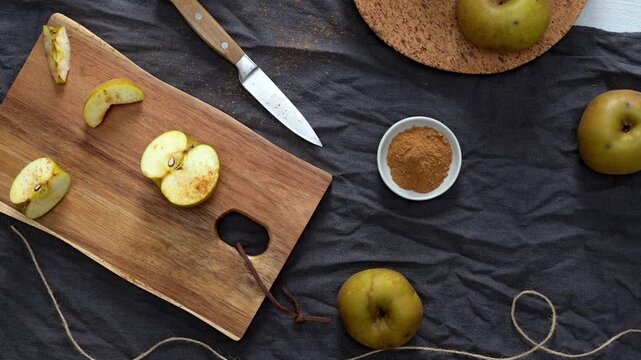 A variety of cut apple slices, dusted with cinnamon, are arranged on a rustic wooden cutting board with a knife, twine, and scissors nearby. The scene, shot from above, captures a cozy, warm mood, hig