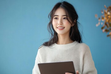 Obraz premium Young woman with long dark hair wearing a white knitted sweater holding a tablet and smiling gently against a blue background
