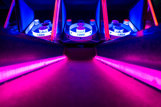 Low angle view of an arcade skee-ball lane illuminated with bright purple and blue neon lights, creating a vibrant gaming and nightlife atmosphere.