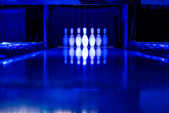 A row of bowling pins illuminated by blue neon lights with a glossy reflective lane surface.