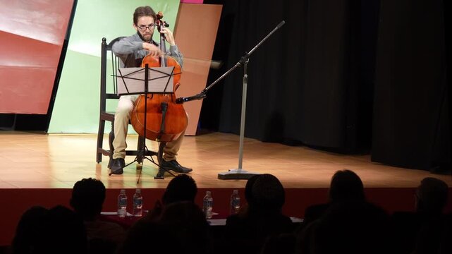 Cellist playing cello performing violoncello solo reading music sheet score on stage with audience in silhouette. Classical music, live performance, artistic expression, string instrument concert