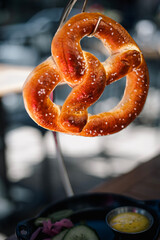 A freshly baked salted pretzel hangs elegantly in front of a blurred background. The pretzel's golden brown color contrasts with the softness of sprinkled salt.
