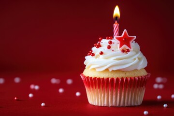Small vanilla cupcake with white frosting decorated with red edible balls and a red star on top with a lit striped candle on a red background