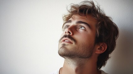 Obraz premium Young man with tousled hair and a beard looking upward against a plain white background, expression thoughtful and curious