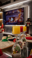 Vertical shot of glowing Christmas candles in festive glass holders, cozy holiday table setting with TV fireplace and seasonal decor.