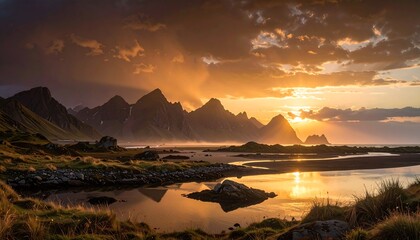 Dramatic sunset paints the jagged peaks of mountains overlooking a tranquil shoreline, showcasing the golden light reflecting on the calm waters.