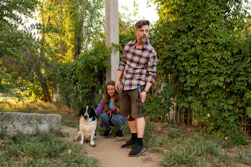 Obraz premium Man stands near his crouching daughter who pets their black and white dog outdoors. Surrounded by greenery, they share a calm family moment in the garden during a warm summer evening