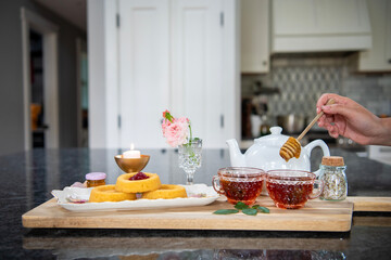 Sweet, slow moment of stirring honey into pink glass tea cups on a tea tray with cakes, jam, and a pink rose. 