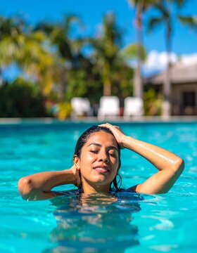 A woman floats peacefully in a turquoise pool, her hands resting on her head. The bright sunny day casts a warm glow, creating a serene outdoor atmosphere.