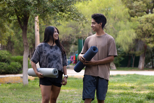 Young couple enjoying conversation in park before yoga session, holding rolled mats, ready for outdoor fitness and healthy lifestyle together