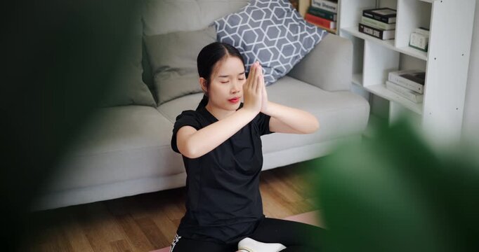 Handheld high angle view shot, Calm Asian woman is enjoying yoga in lotus position on a yoga mat in living room at home. Healthy Lifestyle Concepts