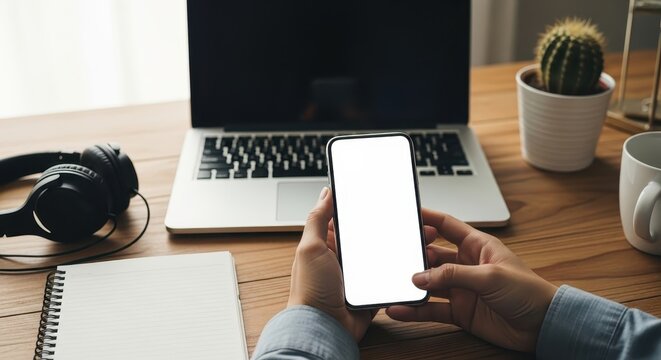 Person holding smartphone with blank screen at workplace with laptop and accessories