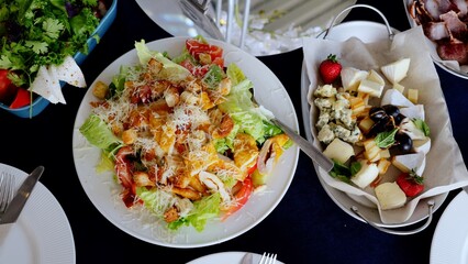 Fresh caesar salad featuring grilled chicken, crispy croutons, parmesan shavings alongside cheese platter, ripe grapes, strawberries on deep blue linen tablecloth