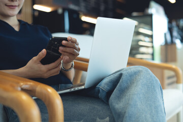 Young asian woman using mobile phone and laptop computer remote working at coffee shop. Woman using...