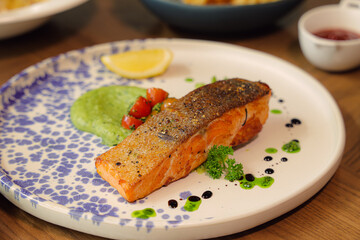 Gourmet Salmon Plate: A close-up view of a gourmet salmon dish on a patterned plate, with expertly cooked salmon fillet alongside vibrant green puree, a fresh lemon slice.