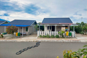 This image shows a row of small permanent houses in a housing complex, featuring blue and black metal roofs, small front yards, and colorful trash bins in front