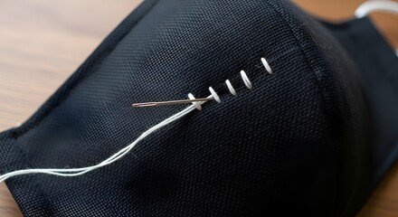 A close-up view captures the intricate process of hand-stitching a protective black fabric face mask with white thread and a sharp needle, highlighting craftsmanship.