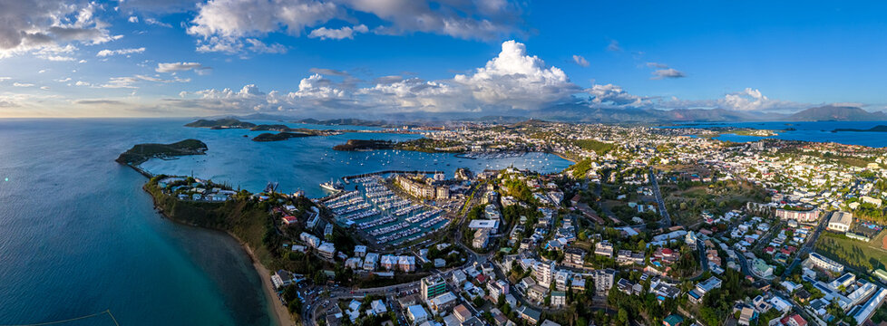 A wide aerial panoramic view of Nouméa, New Caledonia on a bright sunny day. The bustling city, Port Moselle marina, and the surrounding turquoise ocean are captured under a blue sky