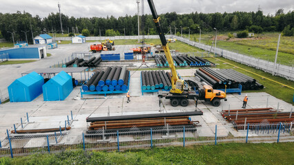 A construction site featuring heavy machinery and workers. Various pipes are stored neatly in rows, surrounded by greenery and industrial buildings