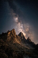A majestic, dark mountain range is silhouetted against a vibrant, star-filled night sky, with the Milky Way galaxy arcing dramatically overhead.  The scene evokes a sense of awe and vastness