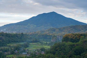A scenic mountain view captured in the morning, showing green forest, rice fields, and a rural village at the foot of the valley in tropical nature.