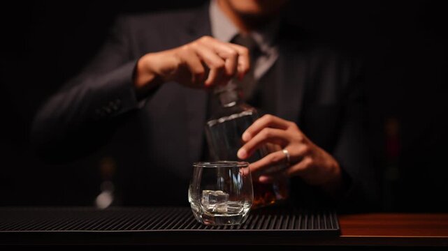 A professional bartender dressed in a stylish red vest and white shirt carefully holds a textured glass whiskey decanter and a glass of amber-colored spirit. The scene is set against a dark, moody bac