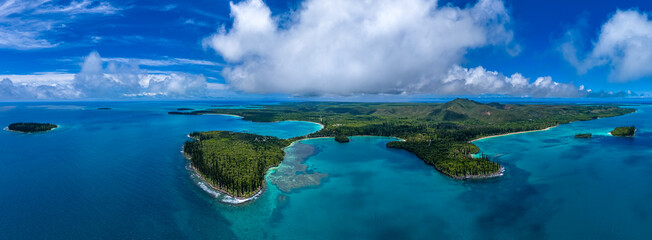 A sweeping aerial panoramic view of the iconic Isle of Pines in New Caledonia. Lush forests of columnar pines cover the landscape, surrounded by the deep blue Pacific Ocean and coral reefs