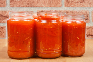 A close-up side view of a stack of glass jars filled with homemade cooked tomato sauce, with a brick wall and wooden surface in the background