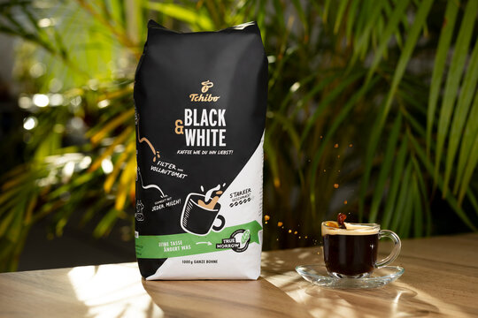 Tchibo Black & White version coffee package on a wooden table with espresso cup and splash action in morning sunlight. Fresh lifestyle concept for coffee culture and brand.