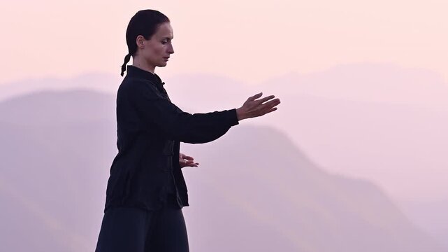 Woman practicing Tai Chi in traditional black outfit on mountain at sunrise, arms outstretched in meditation pose, focusing on balance, energy, and mindfulness in nature.