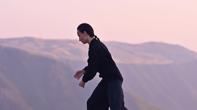 Woman practicing Tai Chi in traditional black outfit on mountain at sunrise, arms outstretched in meditation pose, focusing on balance, energy, and mindfulness in nature.