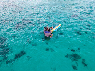 Turquoise ocean scene featuring a small boat and banana boat with several tourists floating over coral formations, captured beautifully from above.