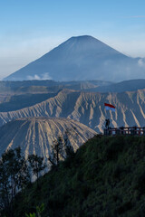 Majestic view of Mount Semeru and Mount Batok with dramatic ridges, a hiker holding the Indonesian flag on a viewpoint, and clear blue sky showcasing Indonesia’s natural beauty.