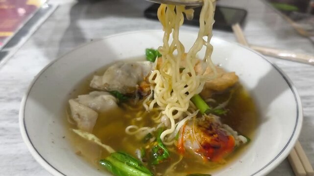 Delicious bowl of Indonesian Bakso with noodles, meatballs, and wontons. A person scoops up the tasty soup.
