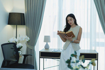Young Asian woman standing by a sheer-curtained window in her home office, engrossed in reading an old book, embodying concepts of learning, knowledge, and quiet contemplation