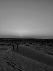 Two children playfully run across the golden sand dunes of Douz, Tunisia, enjoying the tranquil evening as the sun sets in the background, creating a serene atmosphere.