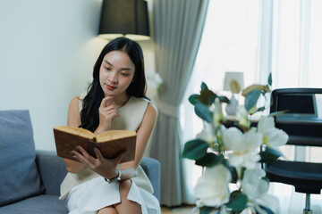 Asian woman reclining on a sofa, absorbed in an old book, enjoying a peaceful, minimalist living room moment of quiet contemplation, comfort and relaxed focus