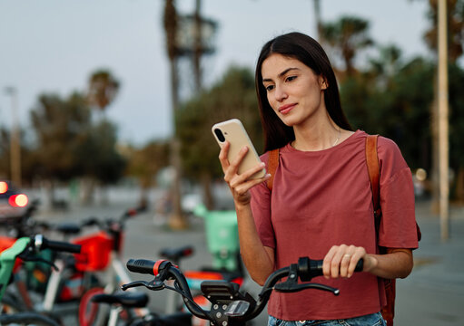 Smiling happy beautiful woman using mobile phone to unlock electric bicycle in city - Powered by Adobe