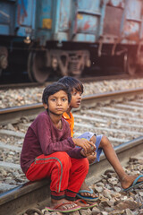 Two Boys Sitting on Railway Tracks Beside Moving Blue Freight Train in Slum Area