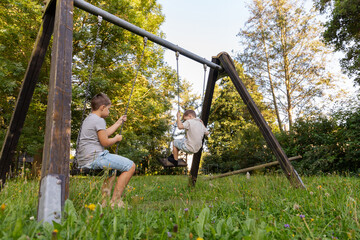 Two kids enjoy playing in a green park, one swinging on a swing set while the other sits nearby, chatting and laughing. Bright sunlight shines through the trees