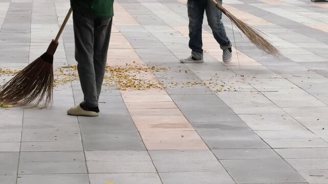 Street cleaners sweeping dry leaves from tiled pavement with traditional brooms