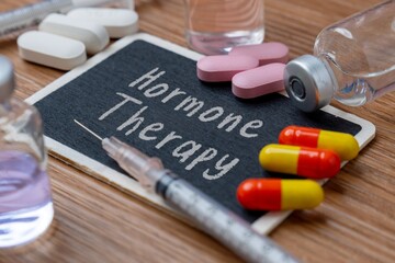 Various pills, capsules, and vials with a syringe on a wooden table. A small chalkboard in the center has the words "Hormone Therapy". Medical and healthcare concept.