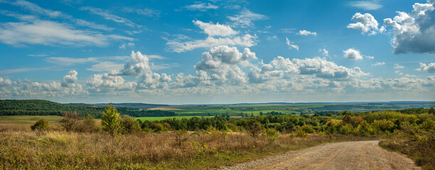 A dirt road leads forward into a landscape with hills, fields and forests under a blue sky