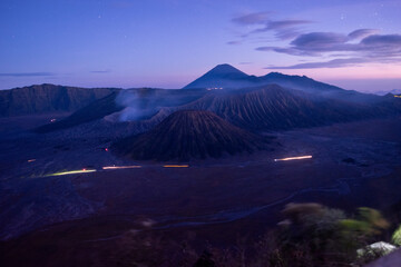 Twilight view of Mount Bromo and surrounding Tengger caldera in East Java, Indonesia, with soft purple skies, misty volcanic slopes, and light trails glowing across the vast desert plain.