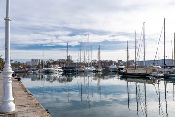 Fototapeta premium Barcelona, Spain-April 12, 2023. A tranquil scene of a sailboats gliding in the harbor with reflections on the still water, surrounded by Barcelona port vell and its nautical vessels