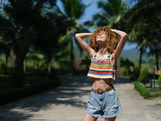 Happy woman enjoying summer outdoors wearing casual colorful top and denim shorts with palm trees and sunny sky background.