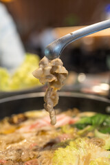 Obraz premium Hands holding chopsticks dipping a slice of pork into a bubbling shabu-shabu pot filled with flavorful broth.